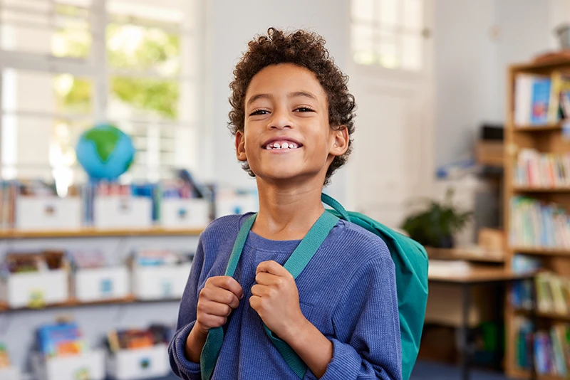 a young boy smiling at school and holding his backpack straps